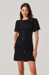 A woman is wearing a short black dress with a fitted silhouette. The dress features short, puffed sleeves with a slight ruffle and is covered in small, shiny sequins that give a sparkly appearance. The neckline is round, and the length of the dress reaches above the knees.